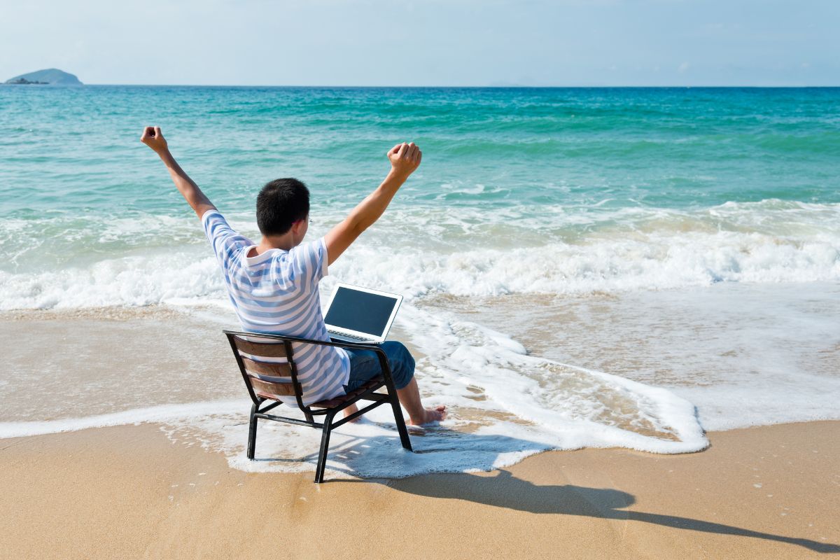 man sitting in a chair raising his hands in the beach with a laptop